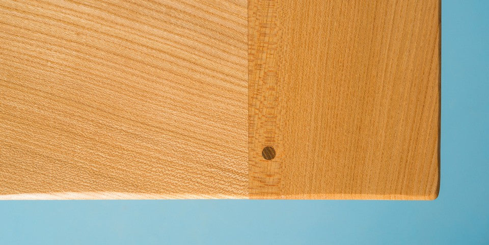 A downward view of the corner of an elm table-top with breadboard ends on a blue background.