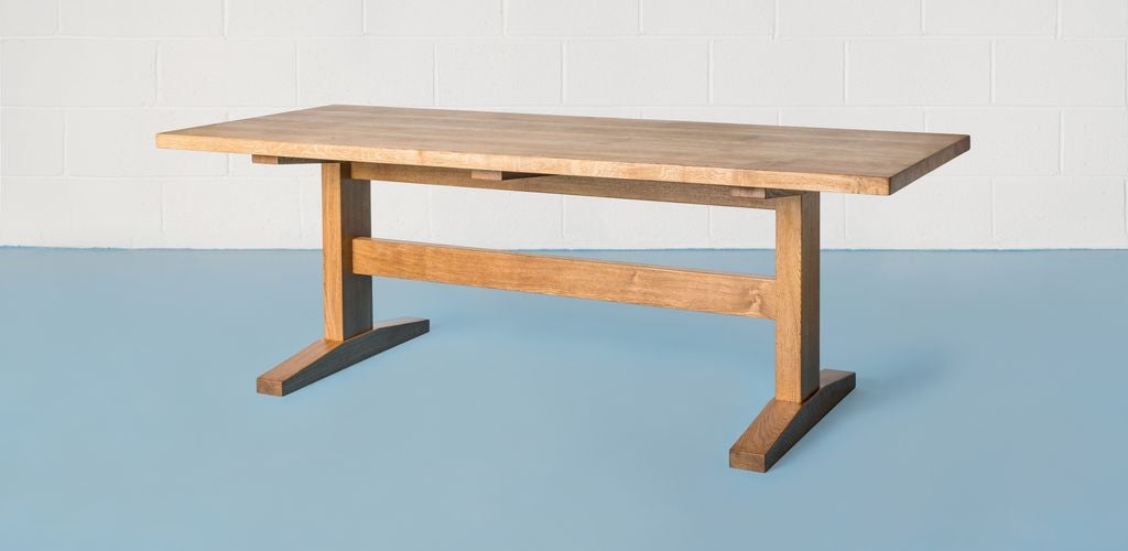 An oak trestle table with an oiled finish stood on a blue floor, with a white blockwork wall in the background.